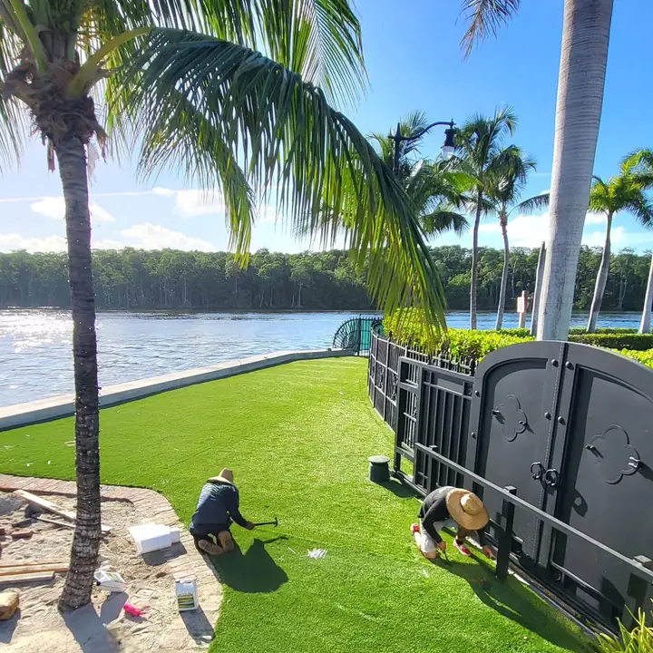 Two installers fastening the edge of new artificial turf along an iron fence beside the water in Miami, Florida