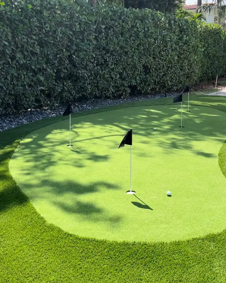 Backyard artificial turf putting green with four flagged cups and a longer fringe collar in front of a clipped hedge in South Florida